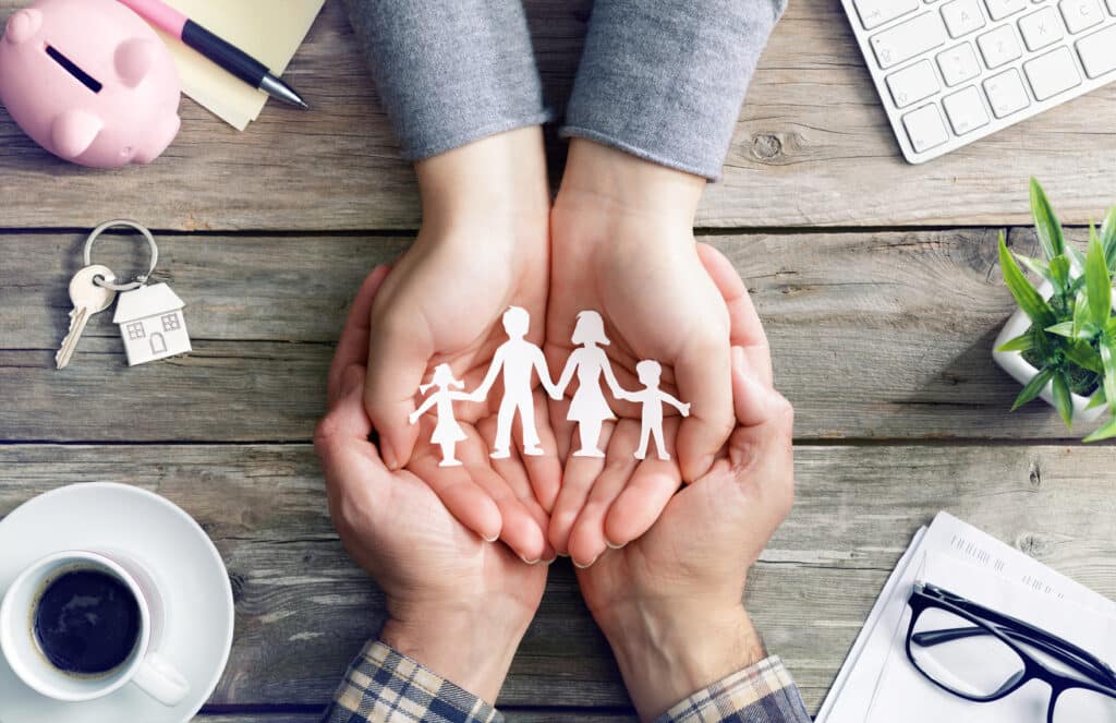 A photo of a male's hands cupped around a woman's hands holding a cutout of a family of four: a daughter, a father, a mother, and a son.