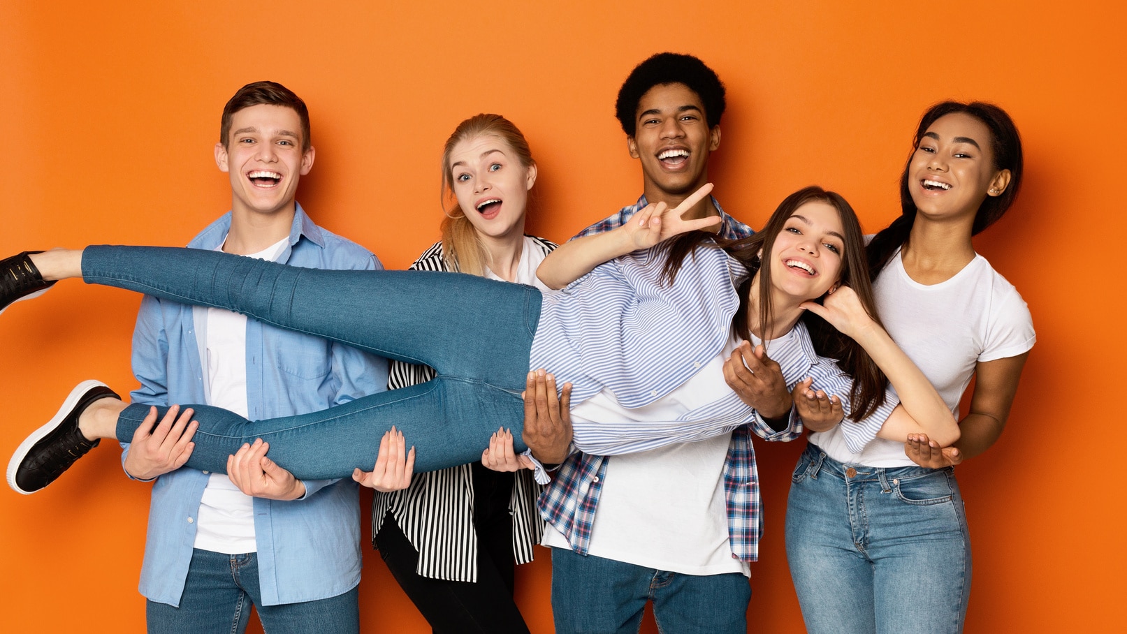 A group of diverse teens posing for a picture while holding up a teenage girl showing off a peace-sign.